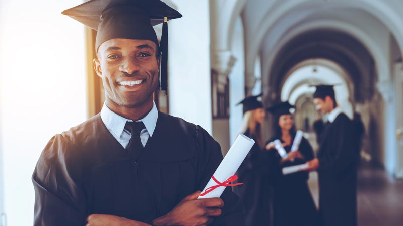 College graduate holding diploma smiling and wearing a graduation cap.